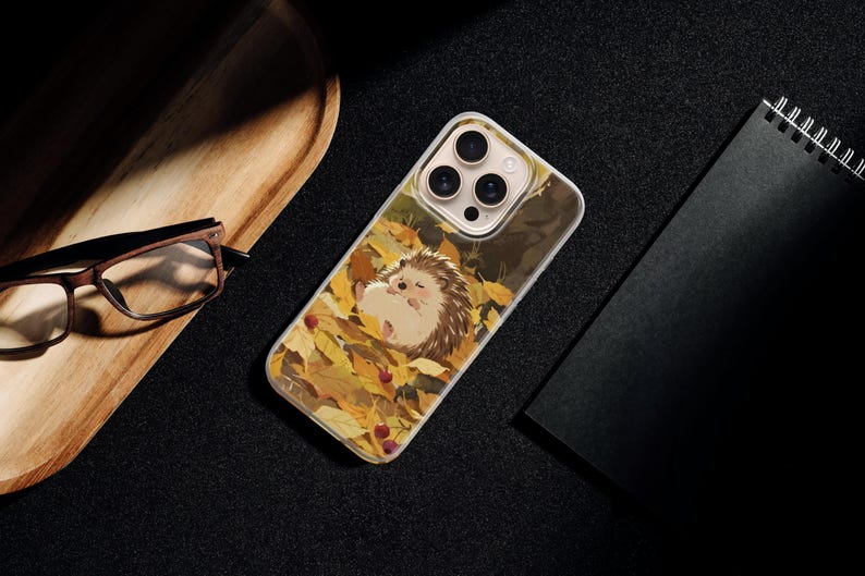 May include: A clear smartphone case featuring a graphic illustration of a small hedgehog sleeping amidst a pile of vibrant yellow and orange autumn leaves, with small red berries scattered throughout the foliage on the back of the device.