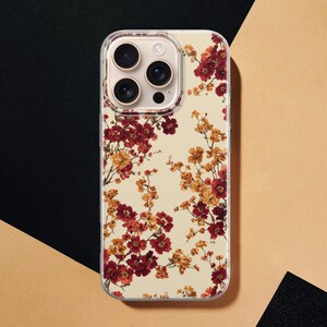 May include: A clear smartphone case featuring a floral pattern with deep red and golden yellow blossoms on thin brown branches. The design is set against a cream-colored background, covering the back of a smartphone with a triple-lens camera system.