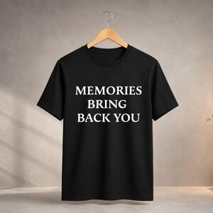 Peut inclure: Un t-shirt noir à manches courtes avec le texte blanc MEMORIES BRING BACK YOU imprimé dans une police sans empattement sur le devant. Le vêtement est présenté sur un cintre en bois devant un fond neutre et texturé.