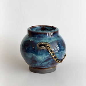 May include: A round ceramic vessel with a glossy, mottled blue and dark brown glaze. The pot features a wide opening, a dark gray base, and a decorative brass-toned metal chain attached to a rectangular metal plate with an engraved pattern.