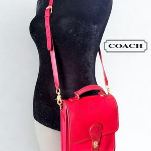 Puede incluir: Un bolso de cuero rojo brillante con asa superior y correa de hombro ajustable. El bolso cuenta con una solapa frontal con cierre de giro dorado y herrajes dorados. La palabra COACH aparece en un marco rectangular a la derecha.