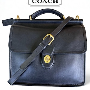 May include: A black leather Coach satchel featuring a top handle, an adjustable shoulder strap with a gold-tone buckle, and a gold-tone turn-lock closure on the front flap. A small black leather Coach hangtag is attached to the side hardware.
