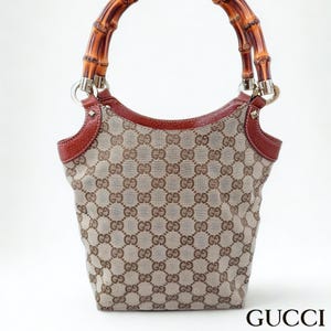May include: A Gucci handbag featuring a beige canvas body with a brown GG monogram pattern, brown leather trim, and a curved bamboo handle. The bag has silver-tone metal hardware connecting the handle to the body. The Gucci logo is visible.