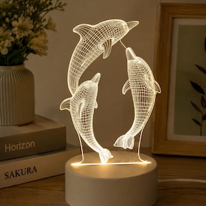 May include: A 3D illusion lamp featuring a clear acrylic panel etched with a wireframe design of three dolphins arranged in a circular formation. The panel is illuminated by a warm white light source housed in a solid, cylindrical white base.