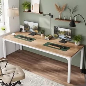 May include: A long rectangular desk with a light wood grain top and white metal legs. The desk surface holds two computer monitors, two keyboards on green desk mats, two computer mice, a stack of books, and a small potted plant.