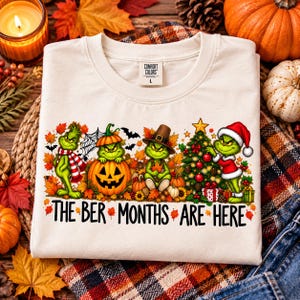 Può includere: Una t-shirt color crema con una grafica del Grinch in vari abiti stagionali, tra cui una zucca, un cappello da pellegrino e un costume da Babbo Natale, circondato da foglie autunnali e un albero di Natale, con il testo THE BER MONTHS ARE HERE.