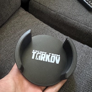 Op de afbeelding: Een cirkelvormige donkergrijze onderzetter met twee gebogen zijwanden, voorzien van de witte tekst Escape From Tarkov in het midden. Het object heeft een matte afwerking en een getextureerd oppervlak, ontworpen om een drankhouder vast te houden.