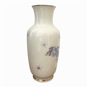 Könnte beinhalten: Eine hohe, cremeweiße Keramikvase mit einer blauen Drachenillustration und goldenen Akzenten. Die Vase hat einen ausgestellten Rand und einen Boden, die beide mit einem dünnen goldenen Metallrand versehen sind. Kleine sternförmige Motive sind verteilt.