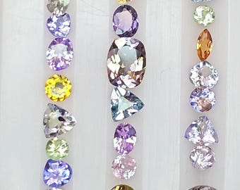 May include: A collection of loose faceted gemstones arranged in three vertical columns on a white display tray. The stones feature various shapes including round, oval, pear, and triangular cuts in a spectrum of colours like yellow, purple, blue, and clear.
