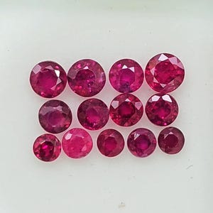 Könnte beinhalten: Eine Ansammlung von dreizehn runden, facettierten, tiefroten Edelsteinen, die in einem Rastermuster in einer weißen Schachtel angeordnet sind. Jeder Stein weist einen Brillantschliff mit zahlreichen Facetten auf, die das Licht reflektieren.