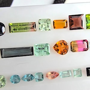 May include: A collection of loose, faceted gemstones arranged in rows on a white surface. The stones feature various shapes including rectangular, square, pear, and round cuts, displaying a spectrum of colors such as green, orange, pink, and blue.