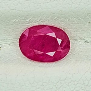 Könnte beinhalten: Ein leuchtender, oval geschliffener Rubin mit einem tiefen, pink-roten Farbton und einer facettierten Oberfläche. Der Edelstein weist eine klare innere Struktur und scharfe, geometrische Facetten auf, die das Licht auf seiner polierten Form reflektieren.