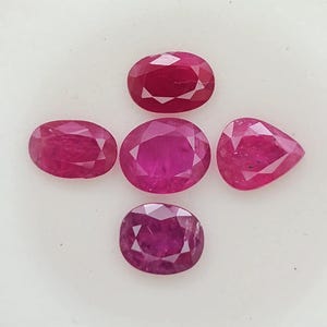 Könnte beinhalten: Fünf facettierte lose Rubine in verschiedenen Formen wie oval, rund und tropfenförmig. Diese Edelsteine zeigen eine tiefe, leuchtende magentarote Farbe mit klaren, geometrischen Schliffmustern auf ihren Oberflächen, kreuzförmig angeordnet.