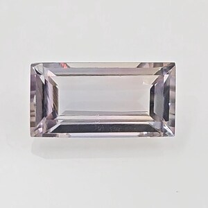 May include: A rectangular, step-cut gemstone with a pale lavender and light purple hue. The stone features a clear, transparent body with sharp, geometric facets that reflect light along its edges, creating a clean, symmetrical appearance.