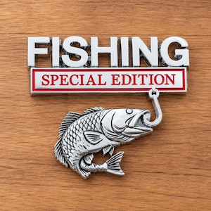 Könnte beinhalten: Ein silberfarbenes Metallemblem mit dem Wort FISHING in fetten Großbuchstaben über einem rechteckigen rot-weißen Rahmen mit dem Text SPECIAL EDITION. Ein silberner Fisch ist unten befestigt und hängt an einem Angelhaken aus Metall.