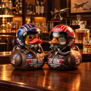 Könnte beinhalten: Zwei Gummienten als Kampfpiloten in braunen Lederjacken mit der Aufschrift Top Gun 40th Anniversary 1986-2026. Eine Ente trägt einen blauen Helm mit der Aufschrift Maverick, die andere einen roten Helm mit Goose, beide mit Visier und Maske.