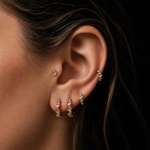 Op de afbeelding: Een set gouden oorringen en een tragus-stud, elk voorzien van kleine, heldere, marquise-geslepen stenen in een clusterpatroon. De sieraden worden gedragen in meerdere piercings langs de oorlel, de helix en de tragus voor een samenhangend geheel.