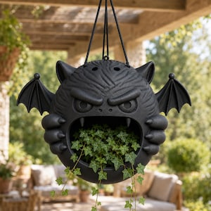 Peut inclure: Une jardinière suspendue noir mat en forme de créature stylisée avec des ailes de chauve-souris, des oreilles pointues et une large bouche ouverte contenant du lierre vert. La jardinière est suspendue par des cordons noirs fixés au sommet.