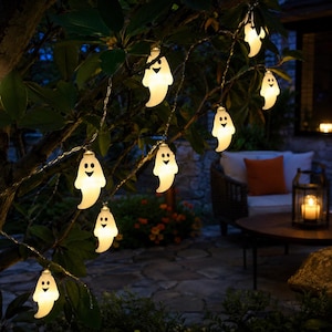 May include: A string of white ghost-shaped LED lights hanging from tree branches. Each ghost features a simple black printed face with two circular eyes and a smiling mouth. The lights emit a soft glow, illuminating the surrounding dark foliage.