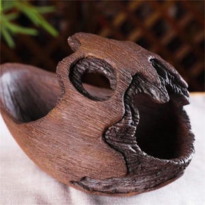 May include: A dark brown, hollowed-out wooden vessel with a natural, rugged exterior texture. The object features an irregular, organic shape with a large, smooth-edged opening on one side and a smaller, circular aperture near the top edge.