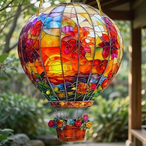 Op de afbeelding: Een heteluchtballon-ornament van glas-in-lood met een levendige zonsondergang in oranje, gele en rode tinten. De ballon is versierd met bloemmotieven en hangt boven een klein mandje gevuld met kleurrijke glazen bloemen.