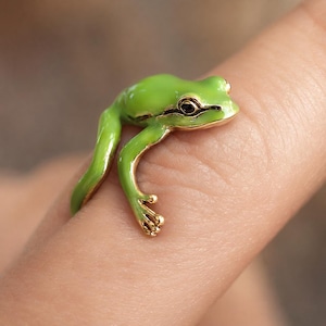 May include: A gold-toned ring shaped like a small frog with bright green enamel detailing on its back and head. The frog design features a dark eye and textured legs that wrap around the finger, creating a realistic and detailed appearance.
