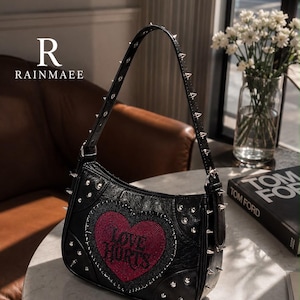 May include: A black shoulder bag featuring a red heart-shaped patch with the text LOVE HURTS in black lettering. The bag is decorated with silver-toned metal studs along the sides and the shoulder strap. The brand name RAINMAEE is visible.