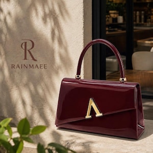 May include: A deep burgundy handbag with a glossy patent finish, featuring a single arched handle with gold-tone hardware and a prominent gold-tone letter A emblem centred on the front flap. The bag has a structured rectangular silhouette.