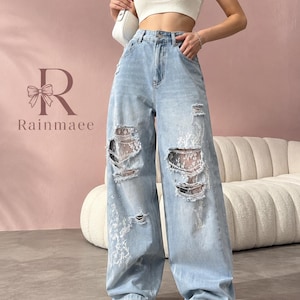 May include: Light blue denim jeans featuring a high-waisted, wide-leg silhouette with distressed cutouts on the front legs. The distressed areas are backed with white floral lace fabric. The jeans include a button and zipper closure.