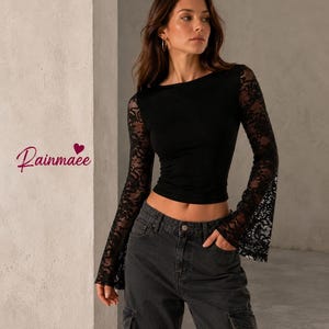 Puede incluir: Un top corto negro de manga larga con mangas de encaje transparente con puños acampanados y un cuerpo negro sólido. El texto Rainmaee con un pequeño icono de corazón es visible a la izquierda. El top se combina con pantalones cargo gris oscuro.
