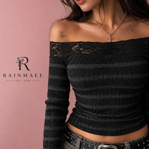 Puede incluir: Una blusa negra de manga larga con textura acanalada y escote tipo barco adornado con encaje negro. La tela presenta rayas horizontales de color gris oscuro. El logo RAINMAEE EST. 2024 aparece a la izquierda. Se lleva un collar con cruz plateada.