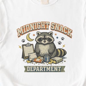 May include: A white t-shirt featuring a graphic of a raccoon surrounded by snacks, a clipboard, and a crescent moon. The text reads MIDNIGHT SNACK DEPARTMENT in orange and green lettering, with small paw prints and lightning bolt accents included.