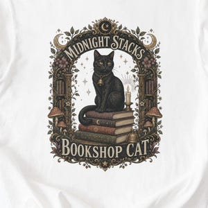 May include: A white t-shirt featuring a graphic print of a black cat sitting on a stack of books. The design includes an ornate frame with mushrooms, a candle, and the text "MIDNIGHT STACKS BOOKSHOP CAT" written in a decorative font style.