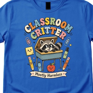 May include: A royal blue t-shirt featuring a graphic of a raccoon inside a school desk. The text reads "CLASSROOM CRITTER" and "Mostly Harmless" on a banner. Surrounding the desk are cartoon school supplies including a pencil, apple, and paper.