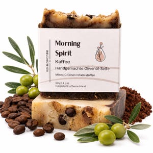 May include: A rectangular bar of handmade olive oil soap labeled Morning Spirit Kaffee with a white paper wrapper. The soap has a marbled brown and cream texture with visible coffee beans. It weighs 90 g / 3.1 oz and is shown with coffee beans.