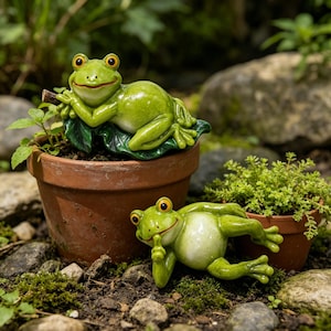 May include: Two glossy green ceramic frog figurines with large orange eyes. One frog rests its chin on a hand while leaning on a leaf inside a terracotta pot. The second frog lies on the ground, holding a finger to its mouth in a shushing gesture.