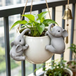 May include: A round, off-white ceramic hanging planter featuring two sculpted grey elephant figures attached to the sides. The planter is suspended by thin twine and contains a green leafy plant with vibrant, heart-shaped foliage.