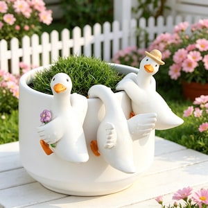May include: A white cylindrical planter featuring three decorative white duck figurines attached to the rim. One duck holds a small purple flower, one leans over the edge, and one wears a tan hat. The planter contains a small green plant.