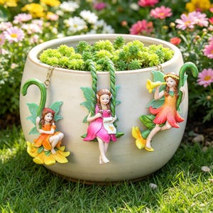 May include: A round, cream-colored ceramic planter filled with bright green succulents. Three small fairy figurines are attached to the rim, each hanging from a green, twisted vine-like hook. The fairies wear orange, pink, and orange outfits.
