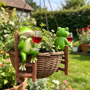 Könnte beinhalten: Ein hängender, brauner Korb aus geflochtenem Material mit grünen Pflanzen und zwei grünen Froschfiguren. Jeder Frosch sitzt auf einer kleinen braunen Leiter am Korb und hält ein durchsichtiges Glas mit einer roten Flüssigkeit in der Hand.