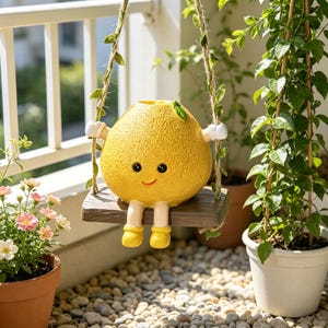 May include: A yellow, textured, citrus-shaped planter with a smiling face, black eyes, and small arms and legs, sitting on a brown wooden swing suspended by two jute ropes. The planter features a small green leaf detail on its top surface.