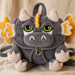 May include: A gray crocheted dragon backpack featuring a plush texture, large green eyes, yellow-accented wings, and light beige horns and claws. The bag includes a silver-toned metal push-lock closure on the front flap and a top handle.
