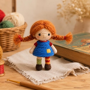 May include: A small crocheted doll with bright orange yarn hair styled in two braids, wearing a blue dress with a yellow patch, one green sock, one striped sock, and brown shoes. The doll has black bead eyes and a stitched smile on its face.