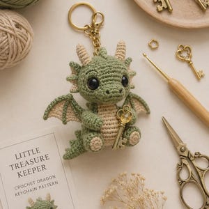 May include: A crocheted green dragon keyring with a cream-coloured belly, wings, and horns, featuring black safety eyes and holding a small gold-toned key. A card labelled LITTLE TREASURE KEEPER CROCHET DRAGON KEYCHAIN PATTERN is visible nearby.