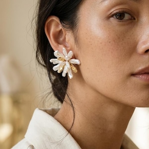 May include: A floral-shaped stud earring featuring a cluster of elongated white baroque pearls interspersed with smooth, polished gold-tone metal accents. The design radiates outward from a central point, creating a textured, multi-layered appearance.