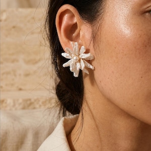 May include: A white floral-shaped earring featuring multiple elongated, irregular petal-like pieces of iridescent baroque pearl radiating from a central, small, smooth, round white pearl bead set on a gold-toned metal base.