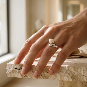 Puede incluir: Un anillo de plata con una banda curva y lisa que se estrecha hacia un diseño abierto, rematado con una perla blanca redonda. El metal tiene un acabado pulido y reflectante que capta la luz, creando un bucle metálico elegante y continuo.