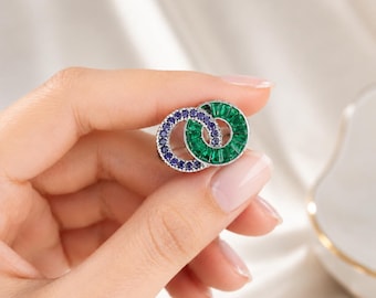 Könnte beinhalten: Eine kreisförmige Brosche mit zwei ineinandergreifenden Ringen. Ein Ring ist mit kleinen runden blauen Steinen besetzt, während der andere Ring mit grünen Baguette-Steinen verziert ist. Die Metallfassung ist silberfarben und bildet einen Kontrast.