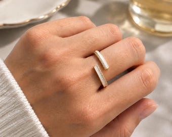 Könnte beinhalten: Ein offener Ring mit zwei parallelen Stäben, die eine weiße Einlage und kleine klare Steine an den Rändern aufweisen. Der Ring besteht aus einem metallischen Material und hat eine Lücke zwischen den beiden Stäben.