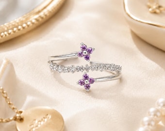 Könnte beinhalten: Ein silberfarbener offener Ring mit einem Mittelband, das mit kleinen klaren Steinen besetzt ist, und zwei Enden in Form von vierblättrigen Blüten, die jeweils mit vier leuchtend violetten Steinen besetzt sind. Filigranes Design.